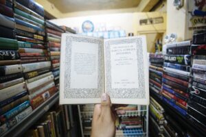 A hand holds an open book with decorative text in a vintage bookstore, surrounded by shelves of books.