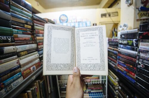 A hand holds an open book with decorative text in a vintage bookstore, surrounded by shelves of books.