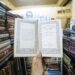 A hand holds an open book with decorative text in a vintage bookstore, surrounded by shelves of books.